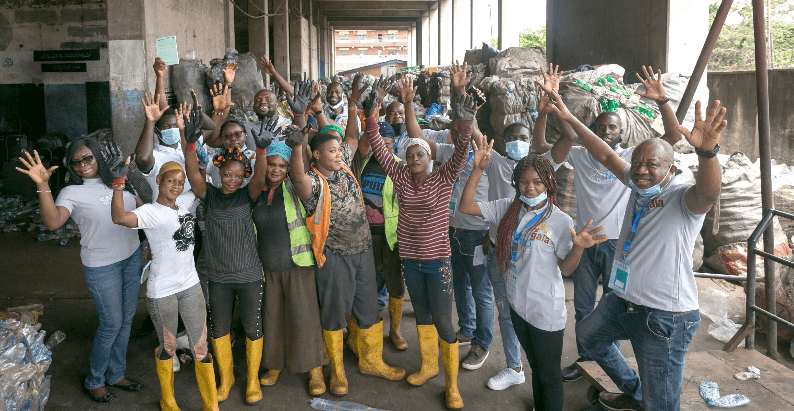 Group of black men and women smiling at camera and raising their arms up in the air. Some of them are wearing yellow rubber boots.