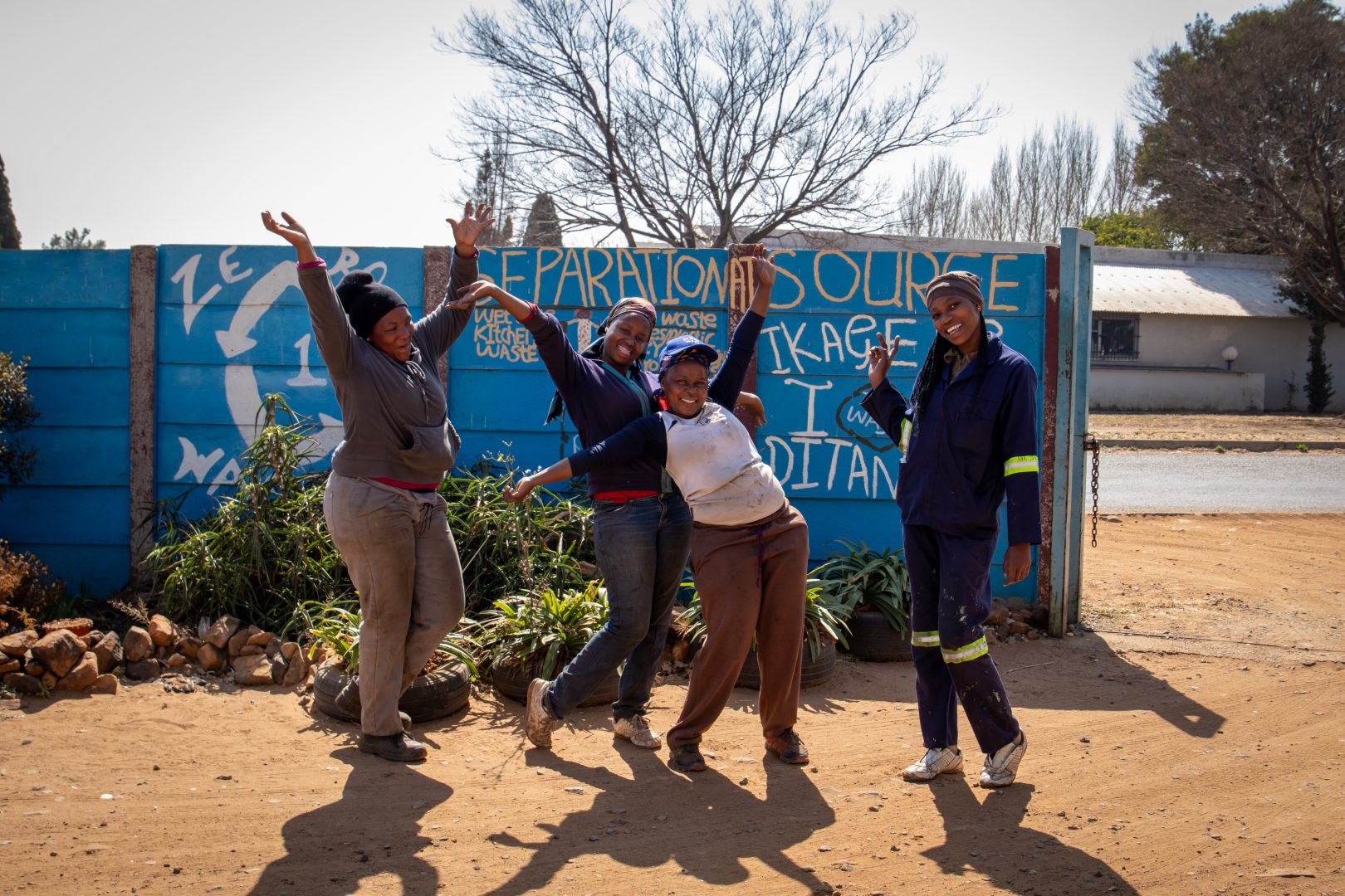Сборщики мусора в Африке весело и беззаботно позируют с поднятыми руками перед синей стеной.