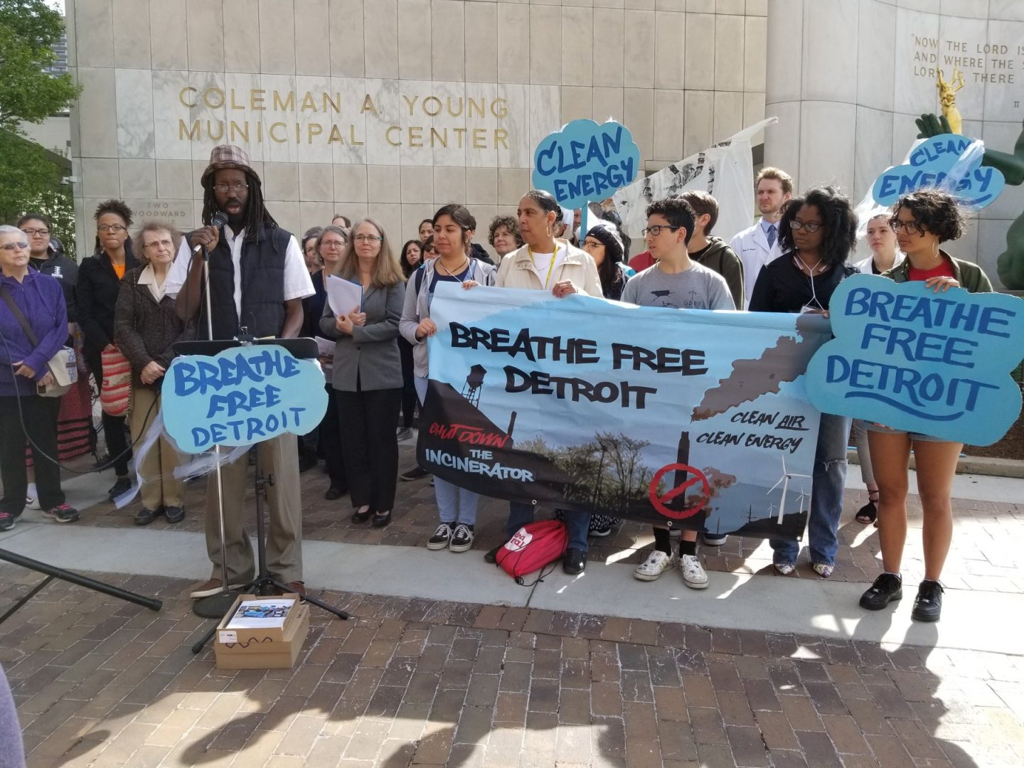 Những người ủng hộ Breathe Free Detroit tập trung xung quanh một người đàn ông cầm micro.