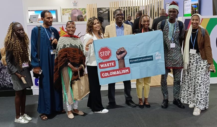 Group of multi racial people standing in front of camera and holding a sign that reads: Stop Waste Colonialism