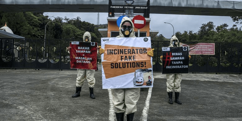 Three protesters wearing complete PPEs protest the incinerator plans in Indonesia. On the left, the protester holds a placard that says in Bahasa Indonesia "Bakar Sampah Masalah Datang", the middle one's placard reads "Incinerator: Fake Solutions!"; and the protester on the right holds a placard that can only be partially seen 